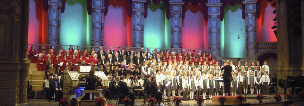 The Vancouver Bach Children’s Chorus in concert on the Orpheum stage: rows of children in white shirts and green ties at the front, adult choristers in red and black behind them, Marisa Gaetanne conducting with her arm raised, the theatre’s ornate arches lit in pink and green and poinsettias along the front of the stage.