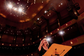 Marisa Gaetanne conducting from the podium, hands raised, in a concert hall lit from above — read more about Marisa